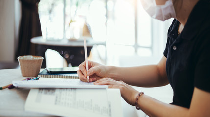 Young asian  woman wearing sanitation mask work and writing on book. she work at home while coronavirus outbreak