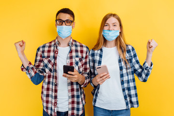 Excited happy guy and girl in plaid shirts and medical masks on their faces, using mobile phones...