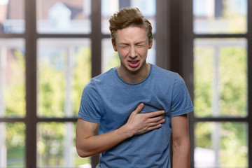 Young caucasian boy suffering from sudden heart attack. Stressed teen guy having chest pain on windows background. Health problem concept. - obrazy, fototapety, plakaty