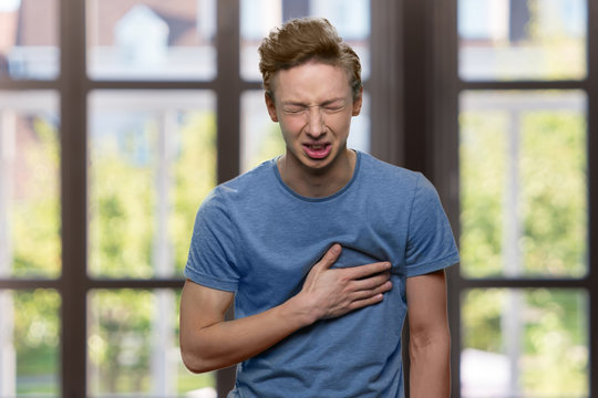 Young Caucasian Boy Suffering From Sudden Heart Attack. Stressed Teen Guy Having Chest Pain On Windows Background. Health Problem Concept.