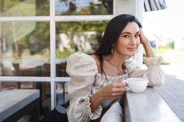 Dreamy attractive woman in fashionable outfit, holding mug coffee, looking camera, enjoying perfect sunny morning, breakfast time. Sexy female at vacation drink cappuccino hotel restaurant