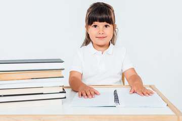 Brunette girl at school, sitting on a chair next to a table with books and a notebook on white background. School concept