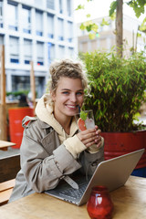 Vertical shot of pretty smiling woman looking happy while drinking cocktail, sitting outdoor table coffee shop, using laptop. Girl studying remote in cafe and enjoying beverage