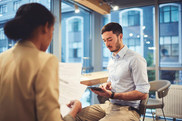 Skilled young male entrepreneur listening attentively to secretary reporting about statistic plannings checking accounting in online database using touchpad connected to wireless internet in office
