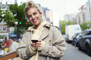 Smiling happy woman standing on street with mobile phone. Beautiful blond girl waiting for cab,...