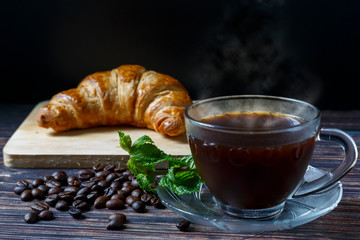 
Hot coffee in a cup placed on a wooden floor with a croissant placed behind it.