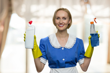 Beautiful house maid holding cleaning supplies and looking at camera. Smiling woman showing two bottles of chemistry for cleaning house. Professional cleaning service.