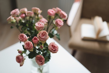 Modern room interior with armchair, book and fresh roses on table. Interior of beautiful living room decorated with flowers