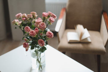 Modern room interior with armchair, book and fresh roses on table. Interior of beautiful living room decorated with flowers