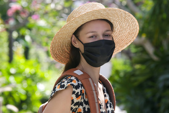 Close up portrait of tourist woman wearing protective face mask. Woman wearing straw hat, medical mask and backpack on her summer vacation. Tourism during coronavirus outbreak.