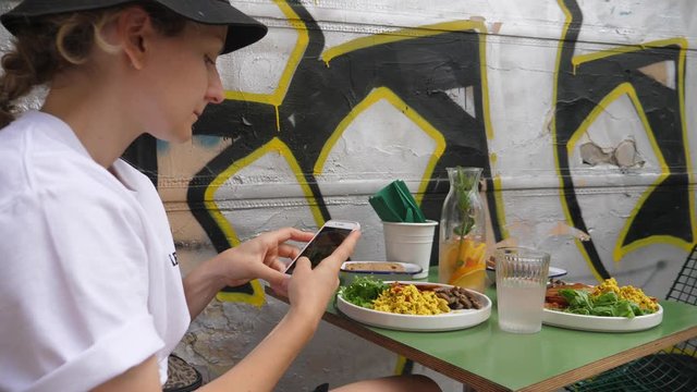 Girl in a bucket hat taking a picture of her breakfast sitting next to graffiti wall.