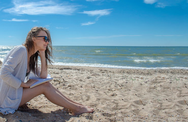 Fototapeta na wymiar Happy woman reads on the beach. European brunette woman in sunglasses reading a book by the sea and looking at the horizon