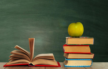 Open book textbook, pile of books and green apple on a wooden desk on the background of a green school chalkboard. Back to school distance home education.Quarantine concept of stay home.