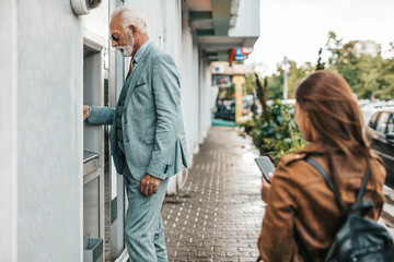 Senior father and his daughter using ATM machine together to withdraw money. They are happy. Bright...