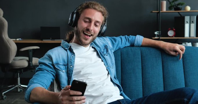 Caucasian Bearded Hipster on the bed and listening to Music in Headphones. Young Man enjoying cool Music on Couch in Loft Living Room. Playfully Mood at Home. FunnyMan. Hobby. Apps.