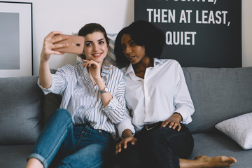 Positive caucasian female blogger shooting video online on front camera of smartphone together with african american friend at home interior.Two hipster girls making selfie on phone sitting on sofa