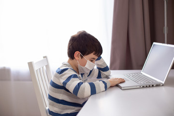 A boy in a white medical mask , sits behind a laptop