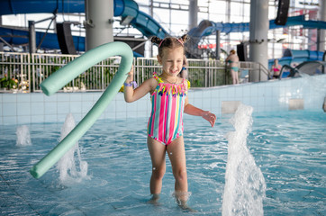 little beautiful girl playing with a foam noodle in a indoor pool. rest in the water park