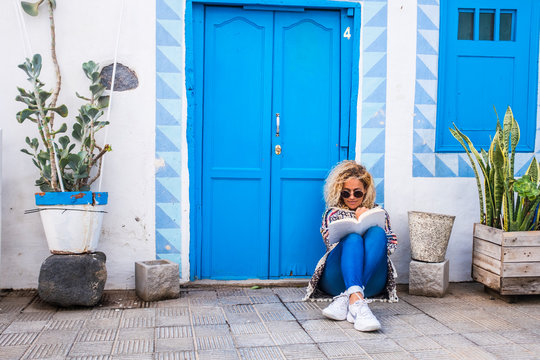 Chererful And Relaxed Beautiful Woman Sit Down Outside A Colorful Blue House In The Street Reading A Book On The Floor - Home Properties Owner People Concept - Travel Lifestyle And Scenic Places 