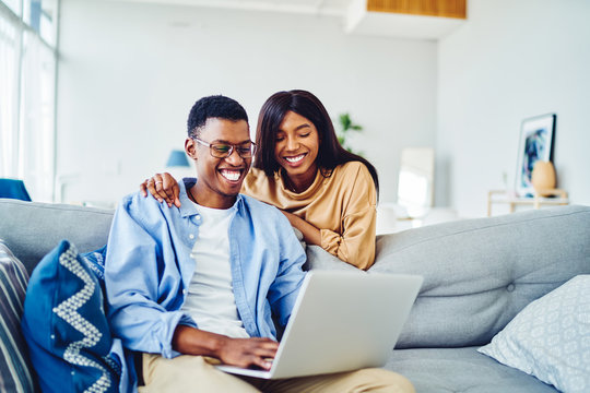 Cheerful african american young marriage laughing while watching movie on laptop computer at home interior, happy dark skinned couple in love viewing funny photos on netbook enjoying free time.