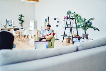 Pensive african american hipster guy reading book during free time in modern designed living room, dark skinned male recreating at apartment with stylish decoration and furniture enjoy literature.