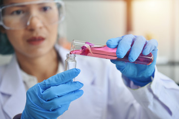 Asian female scientist chemist wearing safety glasses and protective gloves adds pink solution to a...