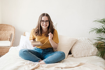 Young beautiful girl student in glasses, an orange T-shirt and blue jeans sits on bed with pillows and reads paper book. Self-education concept at home during quarantine. Home schooling, hobby.