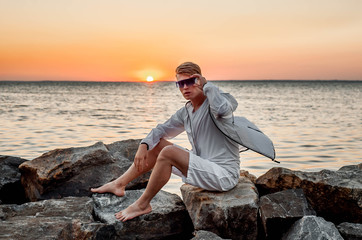 a guy in white clothes and sunglasses sitting on the stones near the sea at sunset