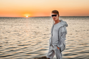 A guy in white clothes and sunglasses stands on the stones by the sea at sunset