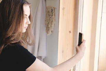 Fototapeta na wymiar Young beautiful woman in a black T-shirt near the window makes a selfie on a smartphone.