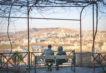 Couple in warm clothes are sitting on the bench and enjoying the view of Holy trinity cathedral  and city view. Tbilisi.Georgia.2020