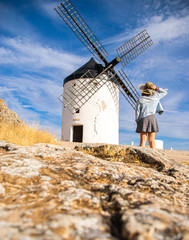 Photo of an attractive woman standing next to some beautiful and historic windmills located in Consuegra, Toledo, Spain during a sunny day of summer in a natural place.  - obrazy, fototapety, plakaty