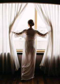 Young woman wearing a robe, looking through the window, holding curtains.