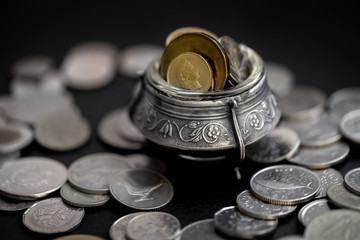 antique silver pot full of gold coins stands among a scattering of silver coins on a dark background