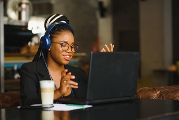 Happy black girl in wireless headphones studying online, using laptop and taking notes, cafe interior, copy space
