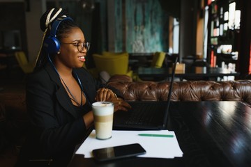 Happy black girl in wireless headphones studying online, using laptop and taking notes, cafe interior, copy space