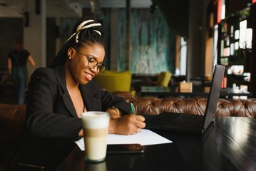 Student girl browsing Internet, using free wi-fi at cafe. African freelancer thinking on ideas for her blog, using laptop at co-working space, resting hand on wooden table, looking with inspired smile