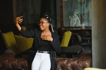 Cheerful dark skinned woman in trendy wear and spectacleson cafe posing for picture and media content for blog, smiling african american female 20s making selfie using mobile phone