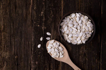 White pumpkin seeds in a bowl, wooden spoon and scattered side by side on a dark wood Image contains copy space
