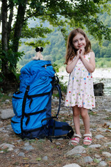 A large backpack for an adult stands next to a little girl against the backdrop of trees and a river, on a sunny day.