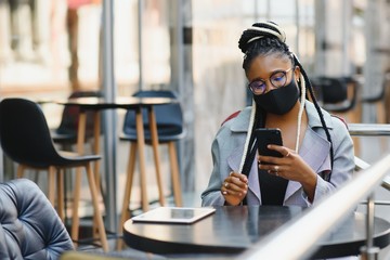 education, healthcare and pandemic concept - african student girl wearing face protective medical mask for protection from virus disease with laptop at cafe