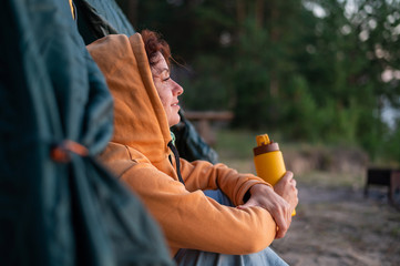 A serene Caucasian woman sits alone in a tourist tent and hold a thermos