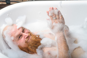 Close-up portrait of funny red-bearded bald man taking a bath with foam. Cheerful naughty guy at...