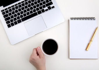  hand holds a cup of coffee on a working light table next to a laptop and accessories. notepad, golden pen. Top view with copy space for text input.  