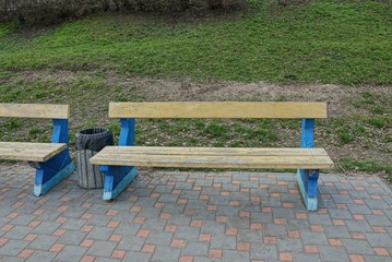 one wooden yellow blue bench stands on the sidewalk near the green grass in the park