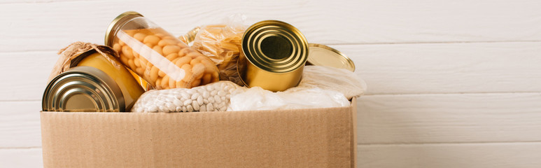 panoramic shot of cardboard box with donated food on wooden background, charity concept