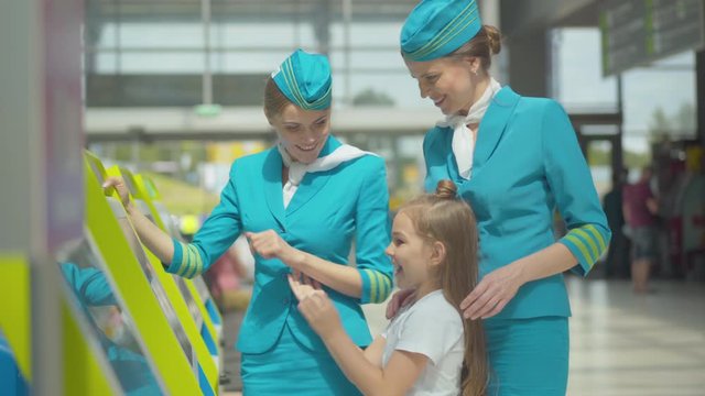 Positive professional stewardesses helping cute little girl with terminal in airport. Side view of pretty Caucasian child making payment supported by smiling flight attendants. Accompanying, tourism.