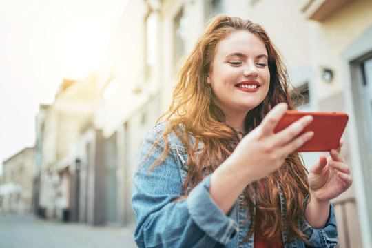 Smiling red curly long hair caucasian teen girl walking on the street and browsing the internet using the modern smartphone. Modern people with technology devices concept image.