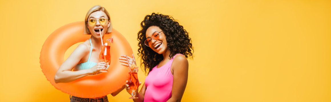 horizontal image of young interracial women with swim ring and cocktail glasses posing isolated on yellow