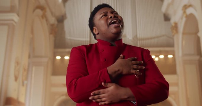 Afro american male preacher singing gospel music. Young man in 30s wearing red suit performing emotionally and moving hands while standing in church. Concept of people and religion